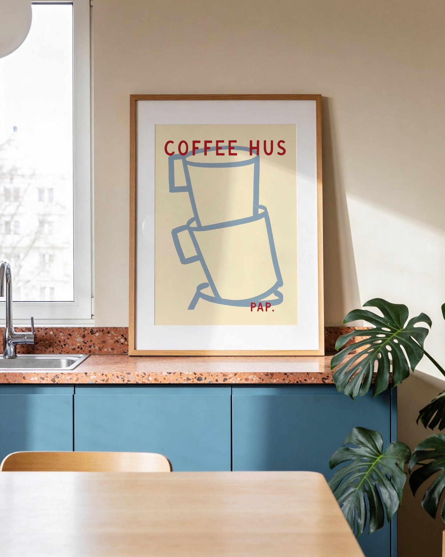 Framed poster of coffee cups on a kitchen wall with a table and plant in the foreground.
